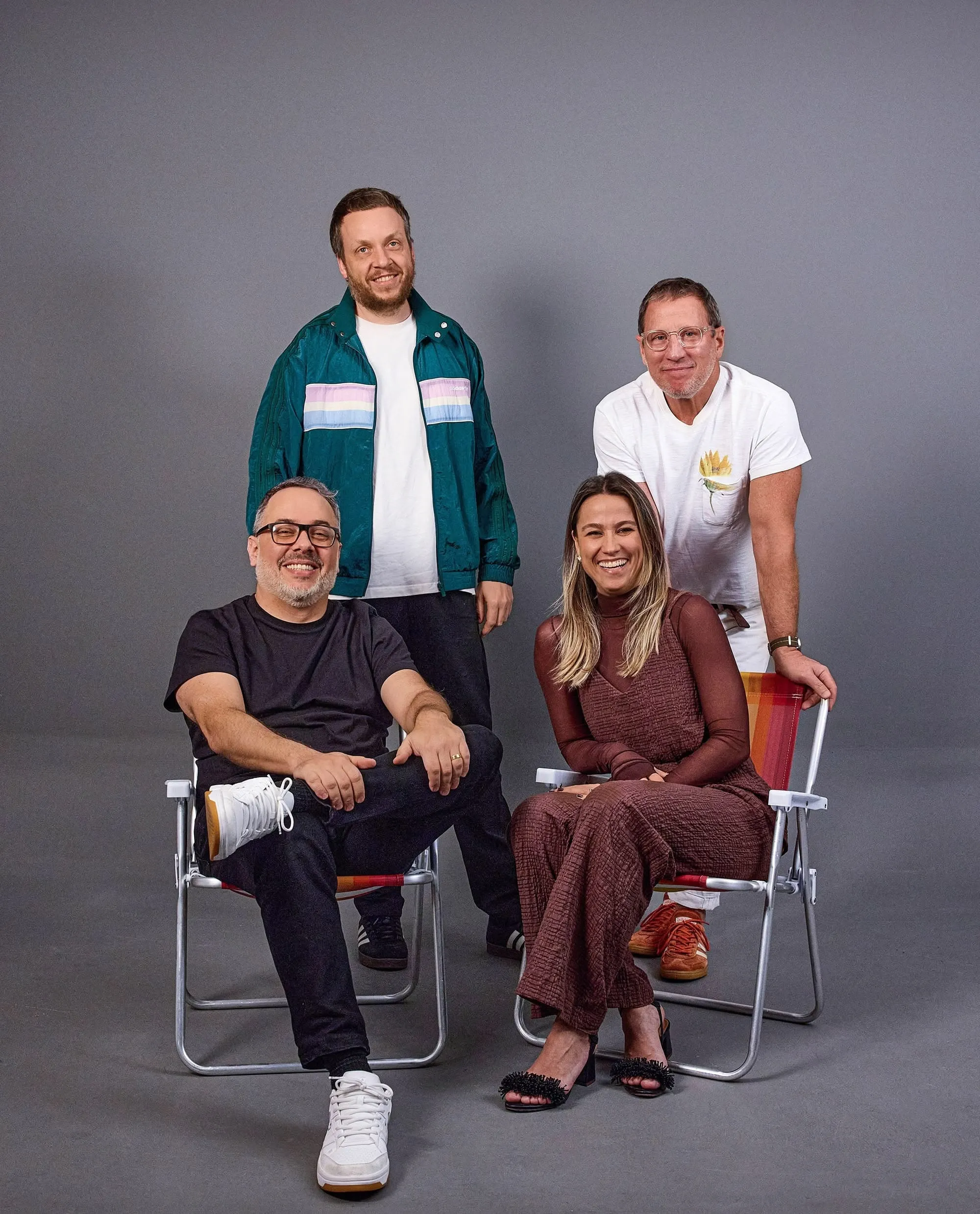 Retrato de estudio del equipo de Praia sobre fondo gris: dos hombres de pie detrás y, al frente, un hombre y una mujer sentados en sillas plegables de playa de aluminio con tela a rayas de colores.