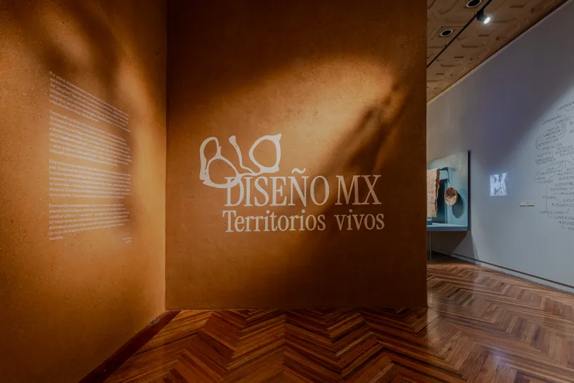 Muro de tierra ocre en la entrada de la exposición Biodiseño MX: Territorios vivos, con el título aplicado en letras claras y sombras de vegetación proyectadas sobre la superficie.