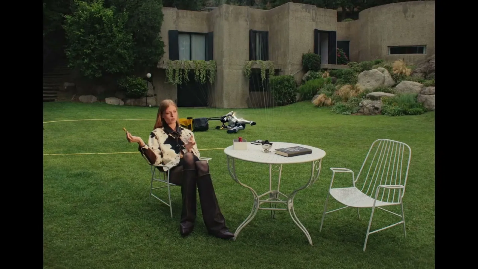 Fotograma de Born to Disobey: Vivian Wilson sentada en una silla de jardín junto a una mesa blanca, mientras el robot DESI 84 yace desarmado sobre el césped entre aspersores encendidos.
