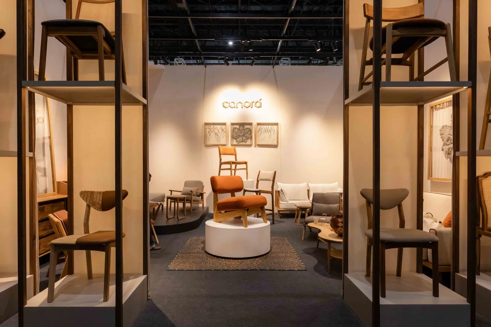 Stand de la marca colombiana Canorá en La Feria de Diseño Medellín: sillas de madera exhibidas en estanterías verticales a los lados y una butaca naranja sobre un plinto blanco al centro.
