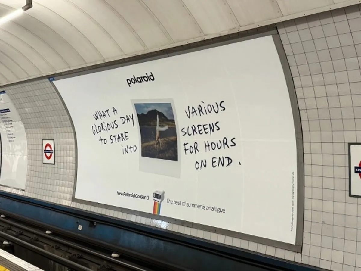 Activación de Polaroid en el metro de Londres, estación King's Cross: un póster sobre fondo blanco con una foto instantánea y el mensaje manuscrito "What a glorious day to stare into various screens for hours on end", con el logo "New Polaroid Go Gen 3 — The best of summer is analogue" y los distintivos del London Underground en las paredes del andén.