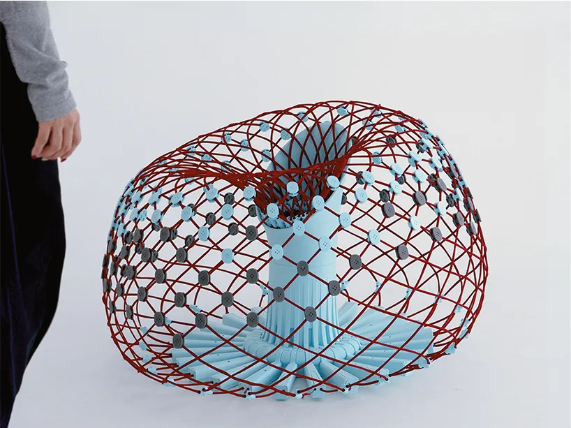 El sofá Re-flat pack desplegado: una estructura tejida con hilos rojos y azules sobre uniones impresas en 3D que forma un asiento redondeado, junto a una persona de pie.