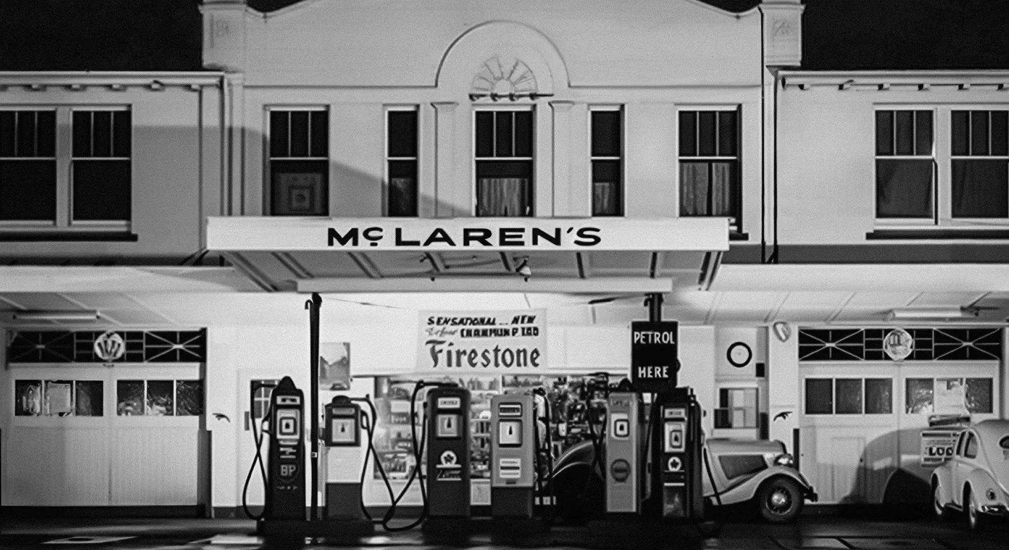 Fotografía en blanco y negro de una estación de servicio de dos pisos con un alero que lleva el letrero McLAREN'S en mayúsculas, surtidores de gasolina antiguos al frente, un cartel de Firestone en la vidriera y dos autos de época estacionados.