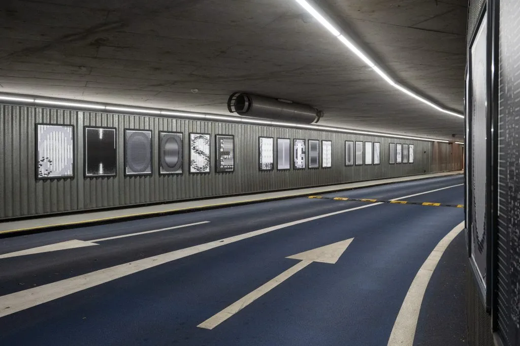 Rampa de estacionamiento de hormigón en Breda con una hilera de pósters monocromáticos enmarcados montados sobre la pared corrugada, iluminados por una tira de luz continua.